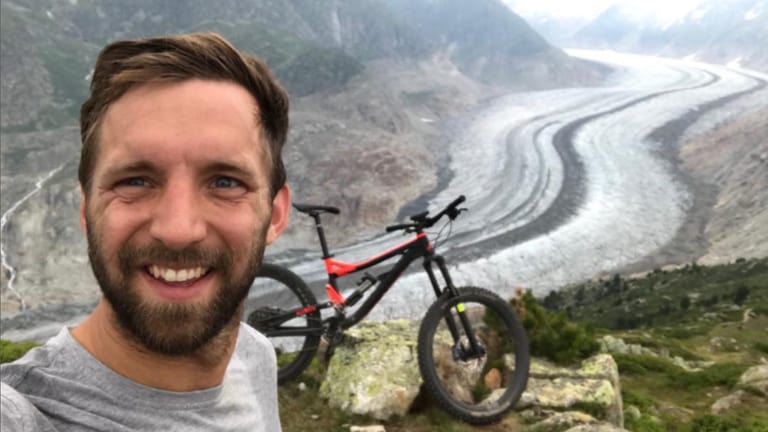 Junger Mann mit braunen Haaren lächelt in die Kamera, im Hintergrund Berge und ein Fahrrad