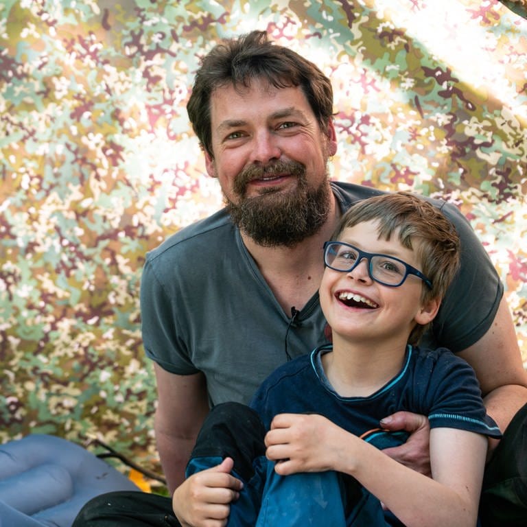 Mann mittleren Alters, Bart und T-Shirt sitzt mit kleinem Jungen im Arm unter einem Zelt (Foto: SWR)