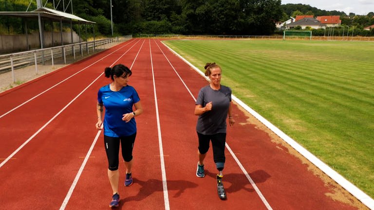 Zwei Frauen joggen auf einem Sportplatz. Die rechte Frau hat eine Beinprothese