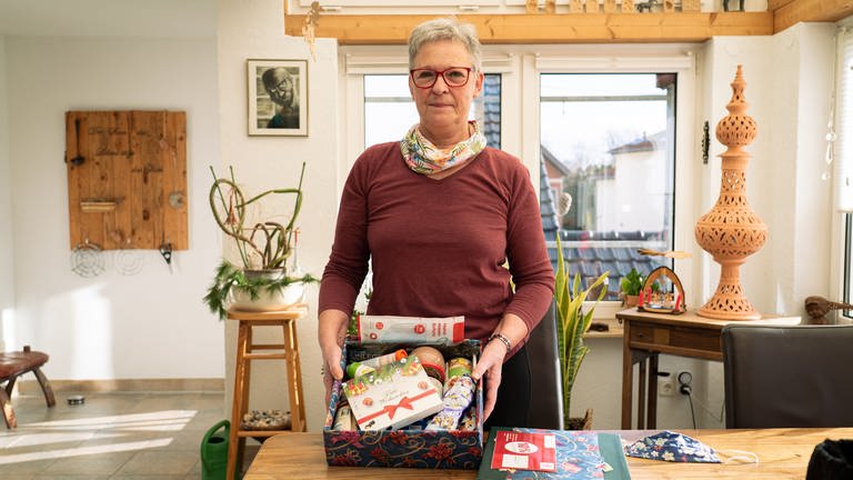 Ursula hat ein weihnachtliches Päckchen in der Hand. In dem Päckchen sind zum Beispiel Schokolade, Kaffee, warme Einlegesohlen und Wurstdosen.