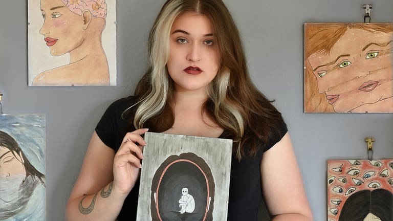 Junge Frau mit einem von ihr gemalten Bild in der Hand, vor einer Wand mit weiteren von ihr gemalten Bildern