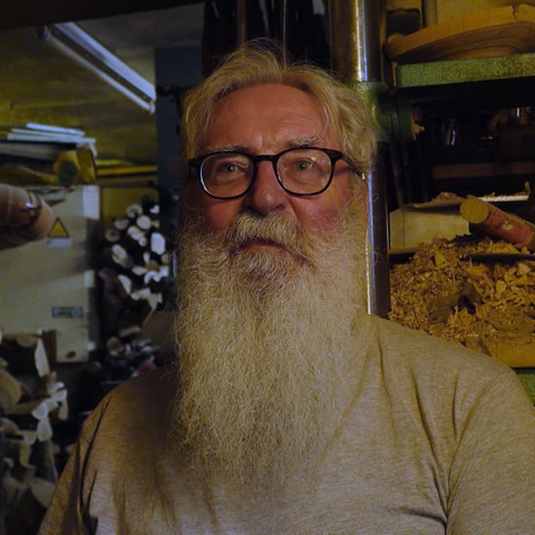 Ein älterer Mann mit Brille und langem weißen Bart steht vor Holzregalen in einem Schuppen